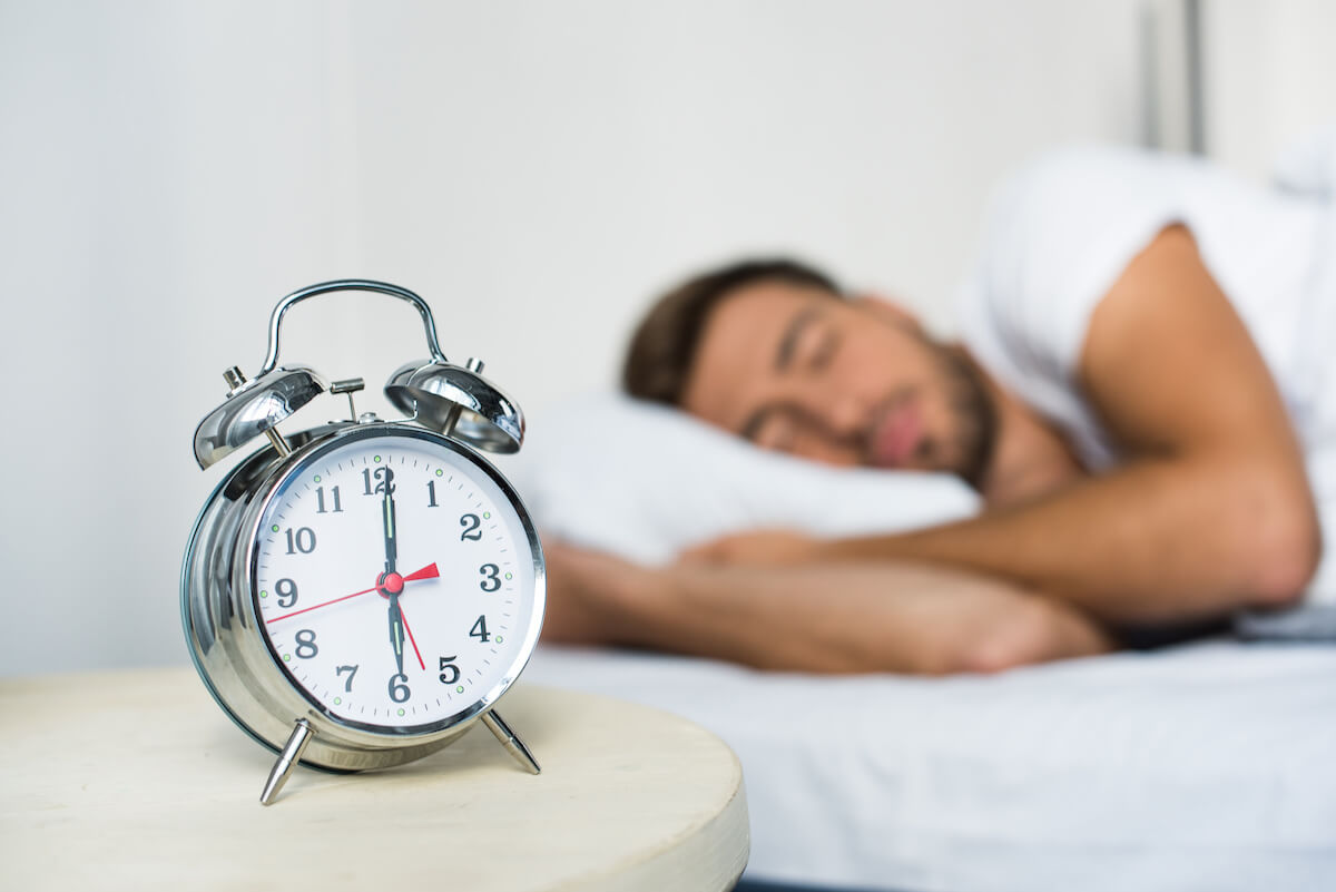 Alarm clock on a bedside table, sleeping man in the background
